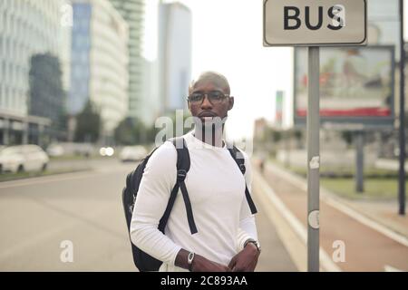 Jeune homme africain attrayant avec des lunettes et un sac à dos debout à l'arrêt de bus à la journée Banque D'Images