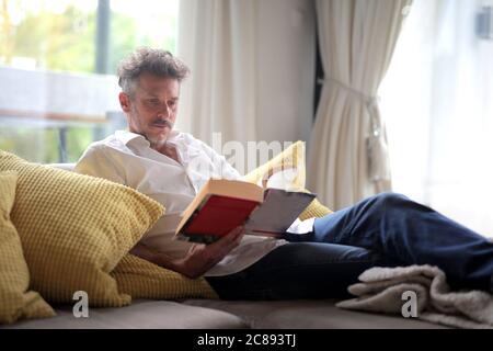 Homme adulte allongé sur le canapé et lisant un livre sous la lumière du soleil par les fenêtres Banque D'Images