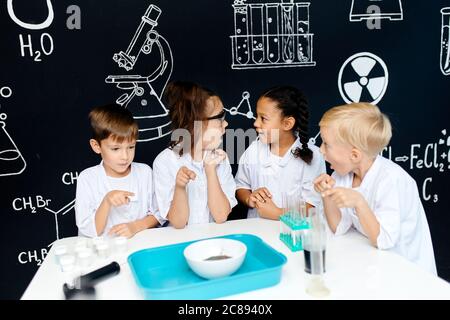 Groupe d'enfants multiraciaux divers dans des vêtements blancs scientifiques faisant des expériences scientifiques avec des réactifs en laboratoire, étant surpris et étonné Banque D'Images