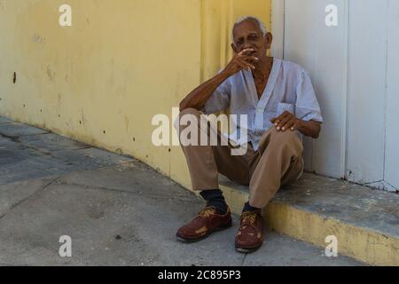 La Havane / Cuba - 04.15.2015: Ancien homme cubain assis dans la rue devant un bâtiment jaune fumant un cigare dans la vieille ville, la Havane, Cuba Banque D'Images