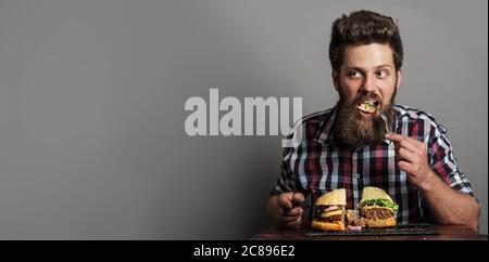 Jeune homme mangeant frais savoureux grand hamburger, fond gris avec espace de copie Banque D'Images