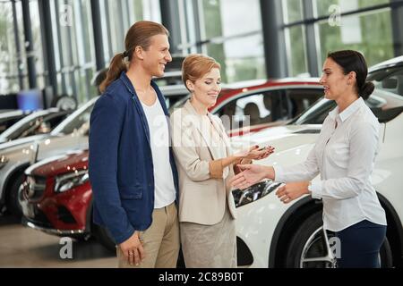 Propriétaires de voitures neuves. Joyeux jeune couple blond habillé de vêtements, recevant les clés d'une voiture d'un vendeur à la concession automobile. Banque D'Images