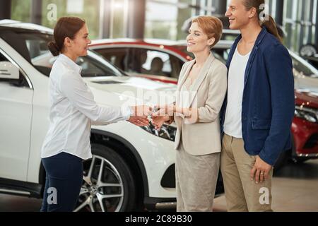 Propriétaires de voitures neuves. Joyeux jeune couple blond habillé de vêtements, recevant les clés d'une voiture d'un vendeur à la concession automobile. Banque D'Images