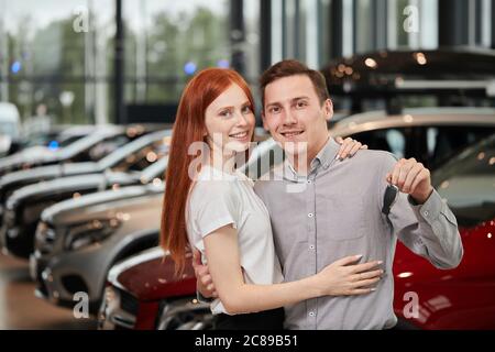 Concept de concession de voitures en visite. Un couple enthousiaste et enthousiaste à la chevelure rouge qui célèbre l'achat d'une nouvelle voiture, tient la clé à portée de main, regarde l'appareil photo et sourit. Banque D'Images