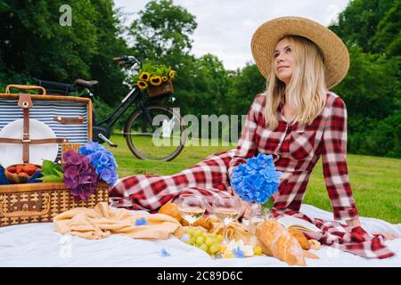 Fille en robe à carreaux rouges et chapeau assis sur une couverture de pique-nique en tricot blanc livre de lecture et de boire du vin. Pique-nique d'été par beau temps avec pain, fruits, bouquet de fleurs d'hortensia. Mise au point sélective Banque D'Images