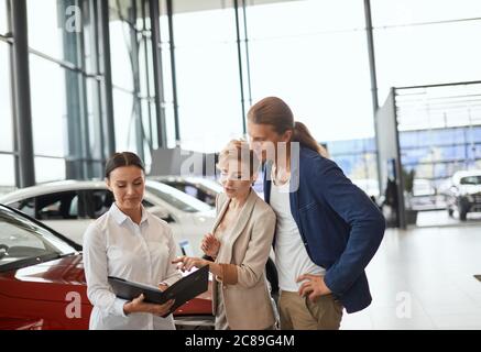 Concept car Business, car concession, clients et commercial. Une femme du chef du service des ventes s'est présentée à un jeune couple dans la salle d'exposition de l'établissement concessionnaire. Banque D'Images