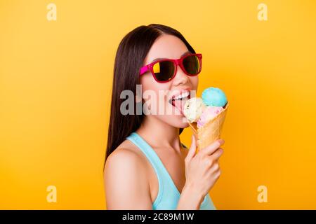 Gaie jeune femme coréenne léche savoureuse glace de trois boules de saveurs différentes, se dresse sur fond jaune en vêtements touristiques, verres Banque D'Images