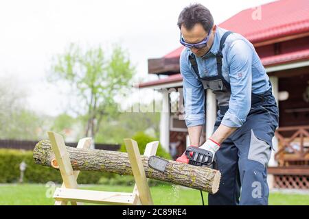 Homme en combinaison scie un arbre sur des sauvettes dans la cour avec une tronçonneuse. Banque D'Images