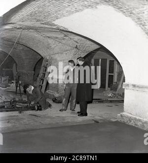 Années 1960, historique, prêtre avec des ouvriers qui entreprennent des réparations à la crypte, à l'église St. Pauls, Deptford, sud-est de Londres. Dans les années 1980, le sous-sol était une discothèque connue sous le nom de 'The Crypt' et le lieu pour les raves et les concerts, y compris le groupe, les Roses de pierre. Banque D'Images