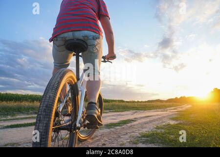 Un gars sur un VTT dans un T-shirt rouge et avec un sac à dos, roule sur une route de terre au coucher du soleil Banque D'Images