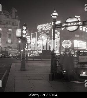 Années 1960, historique, vue sur Piccadilly Circus, Westminster, Londres, Angleterre, Royaume-Uni, montrant les célèbres panneaux d'affichage au néon ou des panneaux publicitaires illuminés sur les bâtiments au rond-point et une entrée au métro de Londres, le métro public de la ville ou le métro. Banque D'Images