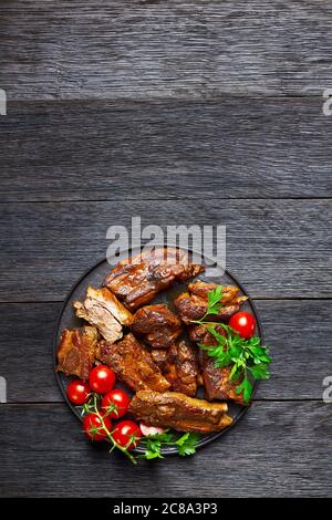 Côtes de porc coréennes épicées lentement cuites avec flocons de poivron rouge, tomates cerises et persil sur une assiette noire sur fond de bois sombre, vue du dessus, Copy spa Banque D'Images