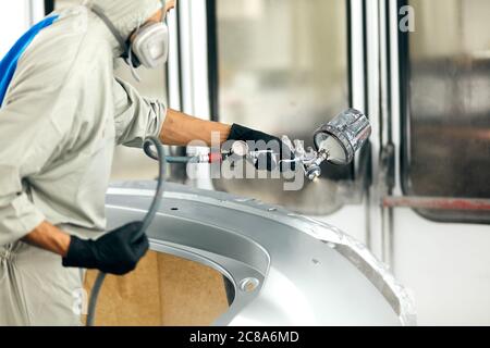 Jeune travailleur peinture des pièces de voiture dans garage spécial, portant costume et matériel de protection Banque D'Images