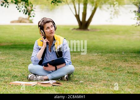 Femme dans un casque assis sur l'herbe verte et étudiant en ligne sur une tablette Banque D'Images