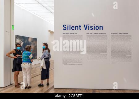 Trois visiteurs portant des masques faciaux discutent devant la grande fresque murale "Silent Vision – Images of Calm and Quiet", qui annonce l'exposition de collection organisée par Raphaël Bouvier à la Fondation Beyeler à Riehen, en Suisse. À gauche, une peinture grand format représentant des nuages sur un ciel bleu est peut-être issue de la série sur les nuages de Gerhard Richter. Banque D'Images