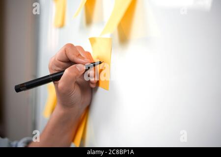 Jeune femme d'affaires du millénaire pensant et écrivant des idées créatives sur des autocollants sur le tableau blanc. Planification du démarrage. Équipe créative travaillant sur la stratégie Banque D'Images