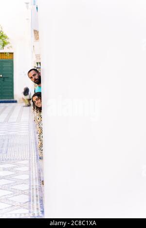 Photo verticale de deux touristes qui collent leur tête de derrière un mur blanc d'un bâtiment au Maroc Banque D'Images