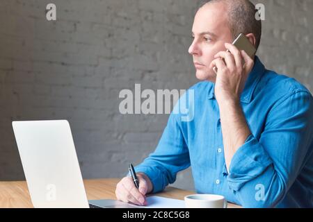 Beau beau beau beau homme mature regarde par la fenêtre et parler sur téléphone cellulaire, assis à l'ordinateur, ordinateur portable. Homme avec des vêtements décontractés en chemise bleue et table Banque D'Images