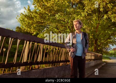 Une femme d'âge moyen se tient sur un pont en face de l'entrée du parc et admire le coucher du soleil. Banque D'Images