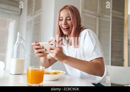 Photo de jeune femme excitée jouant en ligne sur le téléphone portable tout en prenant le petit déjeuner dans la cuisine blanche Banque D'Images