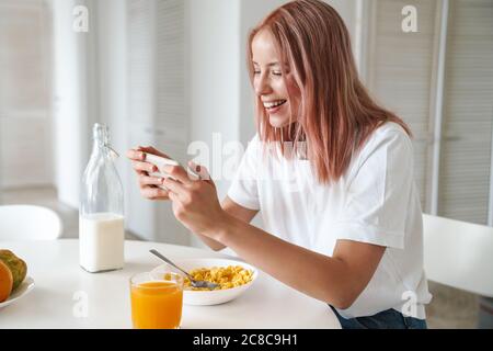 Photo de jeune femme excitée jouant en ligne sur le téléphone portable tout en prenant le petit déjeuner dans la cuisine blanche Banque D'Images