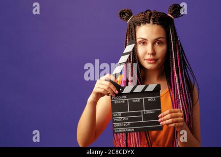 Jeune femme avec des tresses tenant un panneau de clapper en gros plan sur fond violet Banque D'Images