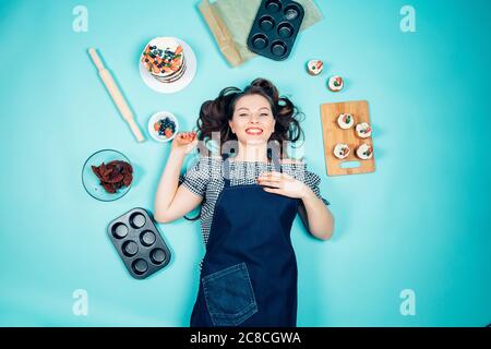 Souriant, le joli chef pâtissier avec des gâteaux et des ingrédients pour cuisiner des gâteaux couchés sur le sol, regardant un appareil photo Banque D'Images