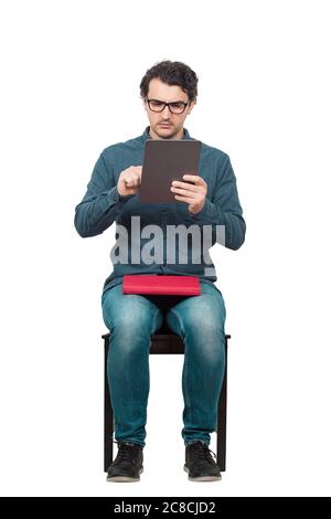 Curieux étudiant homme assis sur une chaise à l'aide d'une tablette PC tout en tient un livre rouge sur ses genoux. Choisissez l'éducation moderne d'ordinateur au lieu de texte traditionnel Banque D'Images