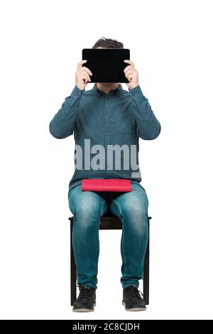 Un jeune étudiant, assis sur une chaise, se couvre de la tête à l'aide d'une tablette PC, tandis qu'il tient un livre rouge sur ses genoux. Concept d'éducation moderne. Étudier et travailler Banque D'Images