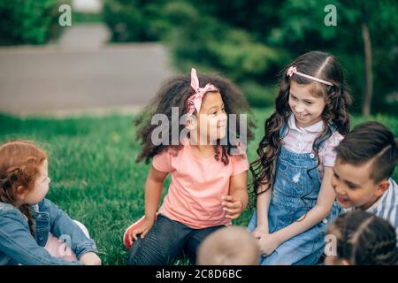 Groupe multi-ethnique enfants assis ensemble souriant à l'extérieur, se concentrer sur les filles africaines. Banque D'Images