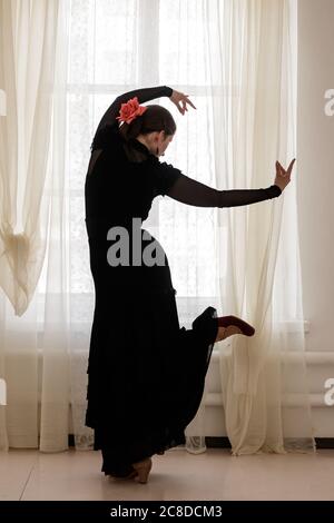 Belle silhouette d'une fille dans une longue robe noire et une rose rouge dans ses cheveux dansant le flamenco sur un fond de fenêtre. Banque D'Images