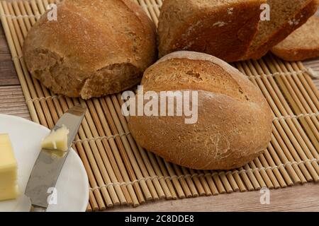 gros plan sur des pains de blé entier faits maison sortis du four. Ils refroidissent sur un tapis de bambou sur une table en bois. Un couteau à beurre avec du beurre. Banque D'Images