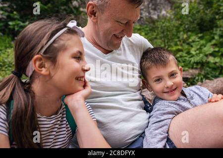 Un père souriant et ses deux enfants adorables assis sur un sentier faisant une pause dans leur randonnée en famille Banque D'Images