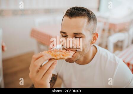 Gros plan du jeune homme avec les yeux fermés manger de la pizza pendant assis dans le restaurant Banque D'Images