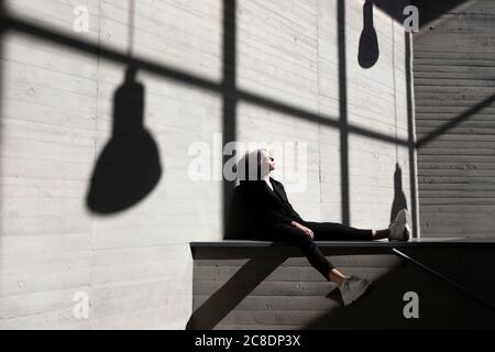 Femme d'affaires vêtue d'un élégant costume assis sur un mur de soutènement exposé à la lumière du soleil et ombre en arrière-plan Banque D'Images