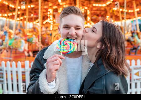 Gros plan de la femme embrassant sur le petit ami cheek mangeant la lollipop à parc d'attractions Banque D'Images