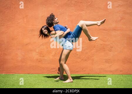 Jeune femme souriante portant l'homme sur le dos par un mur brun par beau temps Banque D'Images