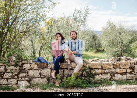 Jeune couple souriant assis sur le mur de soutènement contre les arbres Banque D'Images