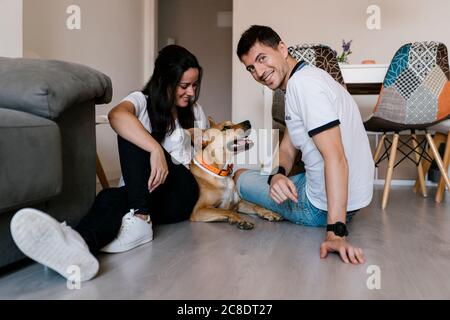 Couple souriant assis avec un chien au sol à la maison Banque D'Images