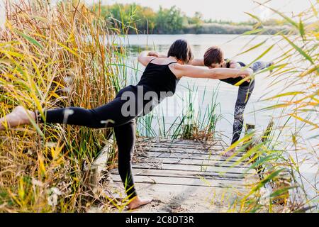 Les amies pratiquant le guerrier 3 posent sur la jetée contre le lac Banque D'Images