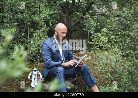 Homme d'affaires chauve utilisant une tablette numérique tout en étant assis avec un robot au milieu arbres en forêt Banque D'Images