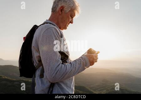 Homme senior utilisant un smartphone pendant la randonnée en montagne pendant coucher de soleil Banque D'Images