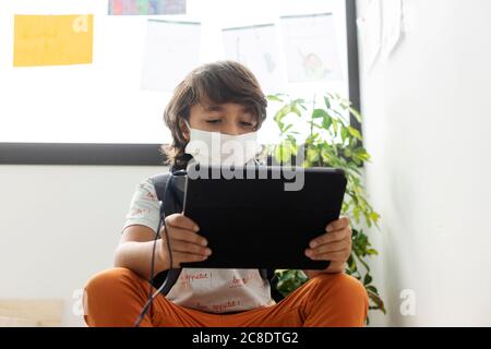 Garçon portant un masque sur une tablette numérique lorsqu'il est assis école Banque D'Images