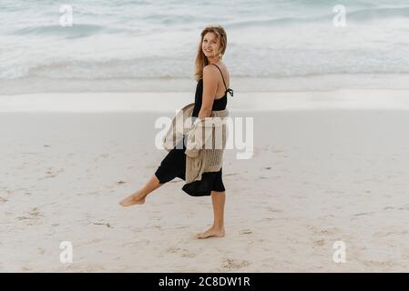 Femme mi-adulte souriante vêtue d'une robe noire marchant à la plage Banque D'Images