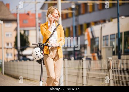 Femme d'affaires utilisant un smartphone debout avec un scooter trottoir en ville Banque D'Images