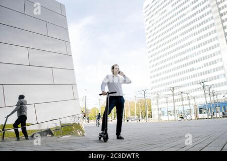 Homme d'affaires avec kick scooter et smartphone se reflète dans la façade du bâtiment Banque D'Images