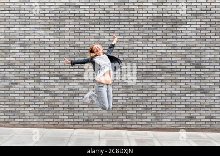 Femme insouciante avec les bras débordés sautant sur le sentier contre le gris mur de briques Banque D'Images
