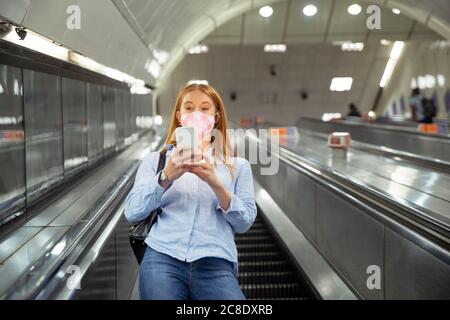 Jeune femme au masque utilisant un smartphone en étant debout escalier roulant à la station de métro Banque D'Images