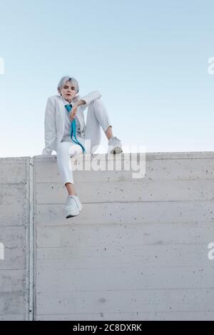 Jeune femme portant un costume blanc assis sur le mur de retenue contre ciel dégagé Banque D'Images