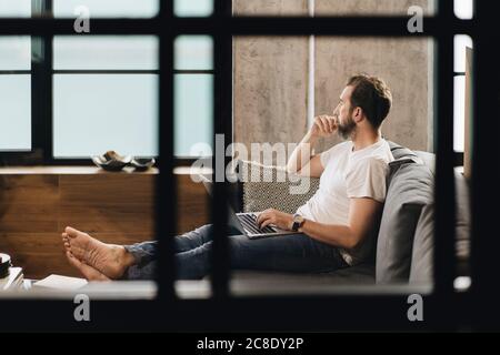 Matur homme assis sur un canapé avec un ordinateur portable sur les genoux, regardant loin, pensant Banque D'Images